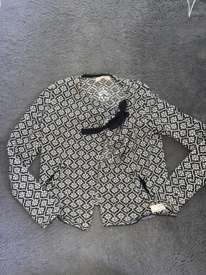 Mönstrad kofta från Odd Molly - Säljer en svartvit mönstrad kofta från Odd Molly med snygg omlottknäppning och dekorativ rosett framtill. Koftan har långa ärmar och ett unikt grafiskt mönster som sticker ut. Perfekt för dig som vill ha något extra till garderoben.