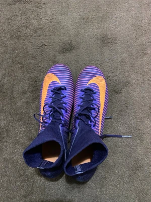 Nike Mercurial lila-orange fotbollsskor - Säljer ett par Nike Mercurial fotbollsskor i lila med orange detaljer och högt skaft. Skorna har snörning, syntetmaterial och dobbar för spel på gräs. Den ikoniska Nike-loggan syns tydligt på sidan och ovandelen. Perfekta för dig som vill sticka ut på planen. Storlek 45 OBS! Möts i Malmö 