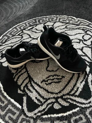 Svarta Nike sneakers med vit sula - Snygga svarta Nike sneakers med vit sula och klassisk vit swoosh på sidan. Skorna har en låg profil och är tillverkade i ett lätt och ventilerande material. Perfekta för en sportig och stilren look.
