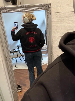 Riktigt cool hoodie/vårjacka från Maje - Cool svart hoodie/vårjacka från Maje med röda detaljer och ett stort rött tigerhuvud broderat med paljetter på ryggen. Väldigt bra skick och alla lappar (och extra knapp) finns kvar. Nypris: 2149.