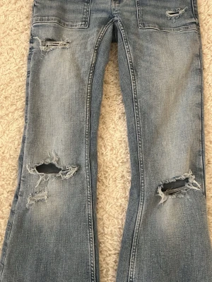 Ljusblå slitna bootcut jeans - Säljer ett par ljusblå bootcut jeans från Ginatricot med slitningar och hål på knäna och låret. Jeansen har två stora fickor fram och dubbla knappar i midjan. Tillverkade i klassisk denim med en cool, avslappnad vibe.Använd endast 3 gånger och nytt pris 400 kr