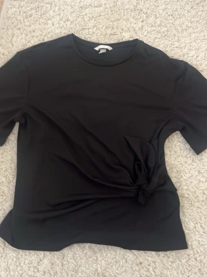 Svart topp med knyt i sidan - Snygg svart topp med rund halsringning och korta ärmar. Toppen har en stilren knyt-detalj i sidan som ger en extra touch till outfiten. Tillverkad i mjukt material som känns skönt mot huden. Perfekt att matcha med både jeans och kjol.