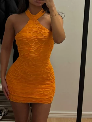 Orange halterneck kortklänning - Säljer en snygg orange kortklänning med halterneck och vågigt strukturmönster. Klänningen är figurnära och har en unik omlott-detalj vid halsen. Perfekt för dig som vill sticka ut med färg och form!
