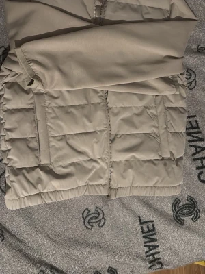 Beige pufferjacka med dragkedja - Snygg beige pufferjacka med raka quiltade paneler och två stora fickor framtill.