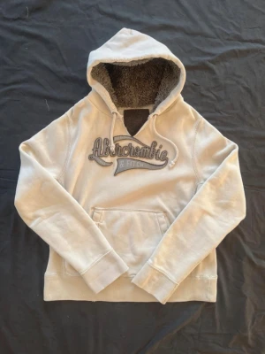 Beige hoodie från Abercrombie & Fitch - Snygg beige hoodie från Abercrombie & Fitch med broderad logga på bröstet. Tillverkad sent 00-tal. Rättså välanvänd och har flera små fläckar (se sista bilderna). Mått (+-1cm) längd 69 bredd 58