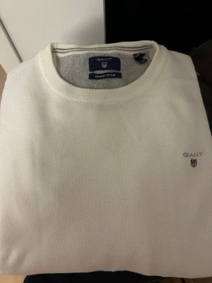 Vit stickad tröja från GANT - Klassisk vit stickad tröja från GANT i premium bomull. Tröjan har rund halsringning, diskret strukturstickning och GANT:s logotyp broderad på bröstet. Perfekt för en clean och stilren look. Storlek M men mer som en S 