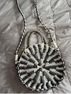 Rund flätad handväska i svart och beige - Säljer en unik rund handväska i flätad design med svart och beige mönster. Väskan har både handtag och en lång axelrem, samt ett rymligt innerfack med dragkedja. Perfekt för dig som vill sticka ut med en bohemisk och trendig accessoar.