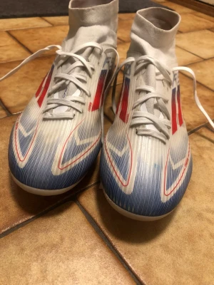 Vita Adidas fotbollsskor med strumpkrage - Säljer ett par vita Adidas fotbollsskor med strumpkrage, röda och blå detaljer samt snörning. Skorna har en modern design med tunna syntetmaterial för bra bollkontroll och grepp på planen. Perfekta för dig som vill sticka ut på fotbollsplanen.