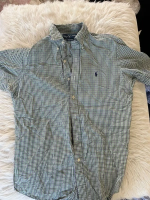 Rutig kortärmad skjorta Ralph Lauren changes - Kortärmad skjorta från Ralph Lauren i klassiskt rutigt mönster i blått och vitt. Skjortan har button-down krage, knappar framtill och en liten broderad logga på bröstet.PIKÉ-känsla i bomull som passar perfekt till sommaren.