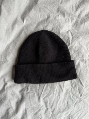 Arket Svart ribbstickad mössa - Säljer en klassisk svart ribbstickad mössa med uppvikt kant. Mössan är tillverkad i en mjuk blandning av 63% ull och 37% polyamid, vilket gör den både varm och bekväm. Perfekt accessoar för kyliga dagar.