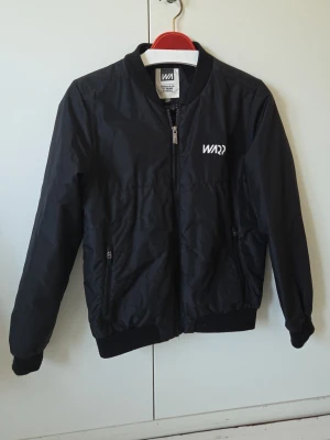 Svart bomberjacka från WARD - Snygg svart bomberjacka från WARP med dragkedja framtill och två fickor med dragkedja. Jackan har ribbade muddar vid ärmslut och nederkant samt en stilren logga på bröstet. Tillverkad i ett lätt och glansigt material som ger en modern look.