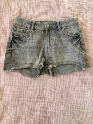 Grå jeansshorts från Lindex - Säljer ett par grå jeansshorts från Lindex med hög midja och råa, fransiga kanter. Shortsen har klassiska fem fickor, knapp och dragkedja framtill samt snygga detaljer på bakfickorna. Perfekta för varma dagar och chill stil.
