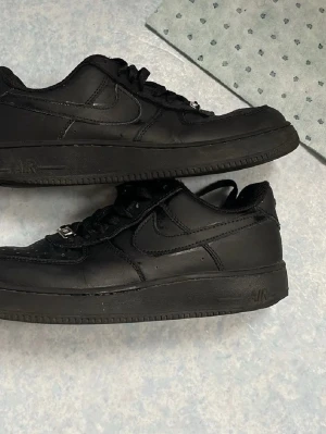 Nike Air Force 1 helsvarta sneakers - Säljer ett par klassiska Nike Air Force 1 sneakers i helsvart läder. Skorna har den ikoniska swoosh-loggan på sidan, svart sula och svarta skosnören. Perfekta för dig som gillar en clean och tidlös look. Passar till det mesta och är riktigt bekväma.