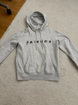 Grå hoodie med FRIENDS-tryck - Säljer en ljusgrå hoodie med svart FRIENDS-tryck på bröstet och klassisk känguruficka. Tröjan har huva med dragsko och ribbade muddar vid ärmslut och nederkant. Perfekt för dig som gillar tv-serien Friends och vill ha en chill stil.