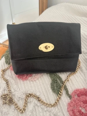 Svart axelväska med guldkedja - Svart, lite glansig, axelväska med guldfärgad kedjerem och vridlås. Har för mig att jag köpte den i Grekland, inte använt den särskilt mycket så därför säljer jag den🤗