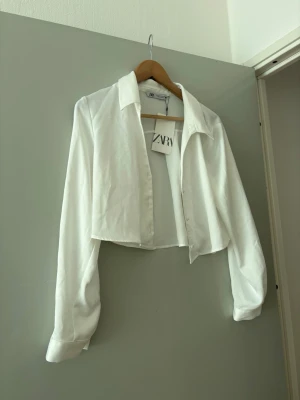 Vit croppad skjorta från Zara - Säljer en vit croppad skjorta från Zara med klassisk krage och långa ärmar. Skjortan har knappar framtill och är tillverkad i ett lätt tyg som ger en fräsch och stilren look. Perfekt för att lyfta vilken outfit som helst.