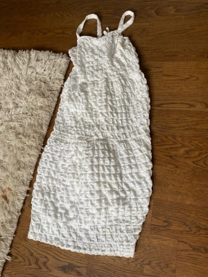 Vit omlottklänning med struktur - Säljer en vit klänning med omlottkänsla och strukturmönster. Klänningen har axelband och är knälång med en lätt volangkant nedtill. Perfekt för sommarens alla tillfällen.