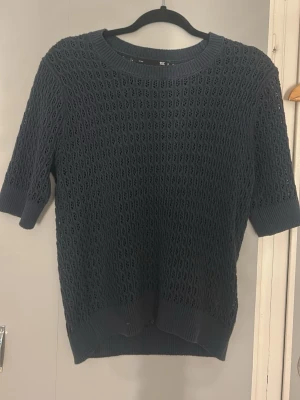 Mörkblå stickad topp med hålmönster - Snygg mörkblå stickad topp med korta ärmar och rund hals. Tröjan har ett genomgående hålmönster som ger en luftig känsla och ribbade kanter vid ärmslut och nederkant. Perfekt för dig som gillar en stilren men ändå unik look.