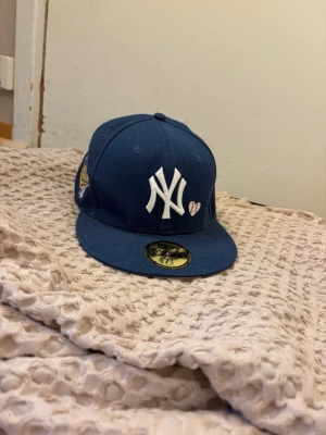 Blå New Era Yankees keps - Säljer en blå New Era 59Fifty keps med New York Yankees-logga i vitt framtill och ett litet broderat hjärta. På sidan finns ett färgglatt World Series-märke. Klassisk rak skärm och broderade ventilationshål. Perfekt för dig som vill ha en sportig och trendig look. Den funkar även super nu när det börjar bli sommar.