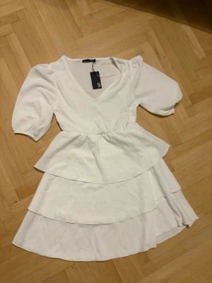 Vit kortklänning med volanger - Supersöt vit kortklänning från lager 157 med puffärmar, v-ringning och dubbla volanglager i kjolen. Klänningen har en feminin siluett och är tillverkad i ett mjukt tyg som känns bekvämt mot huden. Perfekt för sommarens alla tillfällen! Aldrig använd 