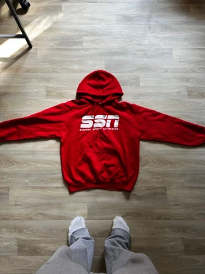 Röd hoodie från SSN - Säljer en röd hoodie från SSN Svensk Sport Nutrition med stor vit logga på bröstet. Hoodien har huva, magficka och ribbade muddar vid ärmslut och nederkant. Perfekt för dig som gillar sportig och avslappnad stil.