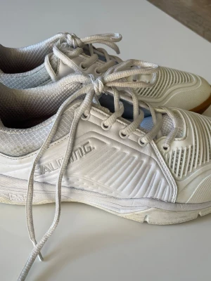 Vita innebandyskor från Salming - Säljer ett par vita innebandyskor från Salming med snörning och gummisula. Skorna har ventilerande meshpartier och detaljer i syntetmaterial. Perfekta för sport eller träning inomhus.