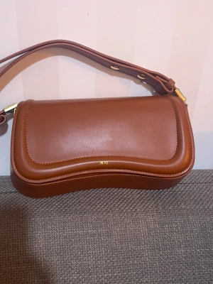 Brun axelremsväska från JW PEI - Snygg brun handväska från JW PEI i veganskt läder med guldfärgade detaljer. Väskan har en unik vågformad design på locket och justerbar axelrem. Magnetlås och ett rymligt huvudfack gör den både praktisk och trendig. Perfekt accessoar för att lyfta din outfit.