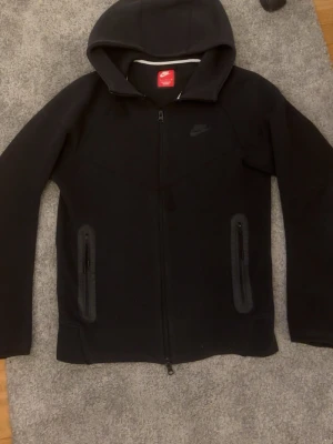 Nike tech hoodie - Säljer en svart hoodie Nike tech med hel dragkedja framtill och två fickor med dragkedja. Klassisk Nike-logga på bröstet och huva. Tillverkad i mjukt material som känns skönt mot huden. Perfekt för dig som gillar sportig och stilren look. Köpt i Göteborg på Nikes egna butik för 1200 och mitt pris är endast 499! Den är använd i väldigt bra skick!🤩😊