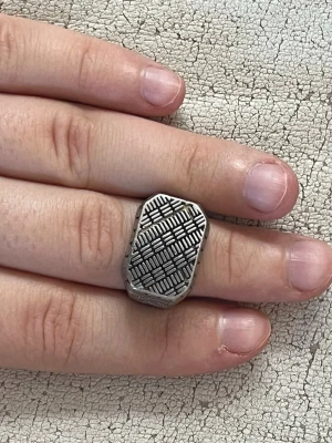 Silvrig ring med mönstrad platta - Unik silvrig ring med en stor, rektangulär platta framtill som har ett detaljerat geometriskt mönster. Sidorna är dekorerade med små hjärtformade detaljer. Perfekt för dig som gillar statement-smycken med cool design.