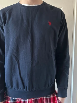 Mörkblå crewneck från Ralph Lauren - Mörkblå sweatshirt från Ralph Lauren med klassisk rund halsringning och röd broderad logga på bröstet. Tröjan har långa ärmar, ribbade muddar och är tillverkad i mjuk bomull. Perfekt för en avslappnad och stilren look.