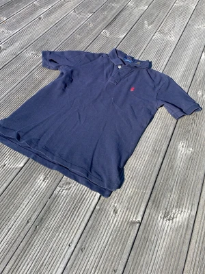 Marinblå pikétröja från Ralph Lauren - Klassisk marinblå pikétröja från Ralph Lauren med korta ärmar och krage. Tröjan har två knappar vid halsen och den ikoniska röda logotypen broderad på bröstet. Perfekt till jeans eller shorts för en avslappnad stil.