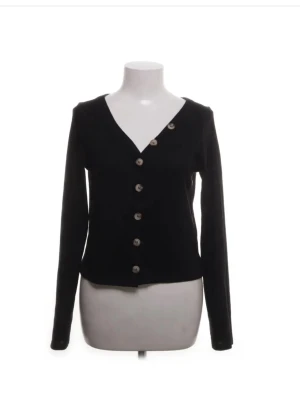 Svart kofta med v-ringning och knappar - Stilren svart kofta med v-ringning och dekorativa knappar längs framsidan. Långärmad modell i mjukt material som passar perfekt till många olika outfits. Enkel men trendig design som är lätt att matcha.