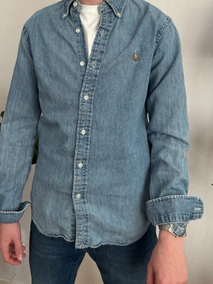 Ljusblå jeansskjorta från Polo Ralph Lauren - Snygg ljusblå jeansskjorta från Polo Ralph Lauren med klassisk krage och knappar framtill. Skjortan har lång ärm och ikonisk broderad logga på bröstet. Perfekt till jeans eller chinos för en avslappnad stil.
