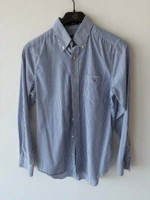 Blårandig skjorta från GANT - Klassisk blårandig skjorta från GANT i modellen The Poplin Banker. Skjortan har vit och blå randigt mönster, knappar framtill och på kragen samt en liten bröstficka med broderad logga. Tillverkad i bomull och har lång ärm.