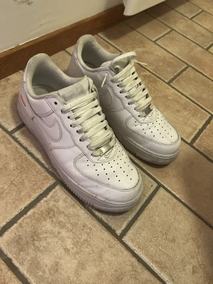 Nike Air Force 1 x Supreme vita sneakers - Klassiska Nike Air Force 1 i samarbete med Supreme. Helt vita sneakers i läder med perforerad tå, vita skosnören och Supreme-logga i rött på sidan. Tidlös design med låg profil och Nike swoosh på båda sidor. Perfekta för dig som gillar streetwear och clean stil.