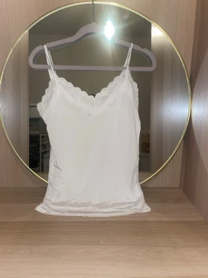 Vit spetsdetaljerad linnetopp - Säljer en vit linnetopp med tunna axelband och v-ringad spetskant i storlek s. Toppen har en mjuk, glansig finish och känns riktigt fräsch och stilren. Perfekt att styla lager på lager eller bära som den är för en clean look.köpt på bikbok och är nästan aldrig använd!skriv för fler bilder,pris kan diskuteras!💗(säljer även en exakt likadan i svart)