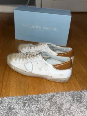 Vita Philippe Model sneakers med bruna detaljer - Snygga vita sneakers från Philippe Model med bruna detaljer vid hälen. Skorna har klassisk låg profil, vita snören och en robust sula. Tillverkade i läder med stilren design och diskret logga på sidan. Perfekta för dig som gillar en clean och trendig look.