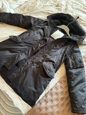 Svart parkas med huva  - Snygg svart parkas med stor huva fodrad med mjuk fuskpäls. Jackan har dubbelknäppning med både dragkedja och knappar, flera fickor framtill och en ärmficka med dragkedja. Perfekt för kalla dagar och riktigt skön att bära.