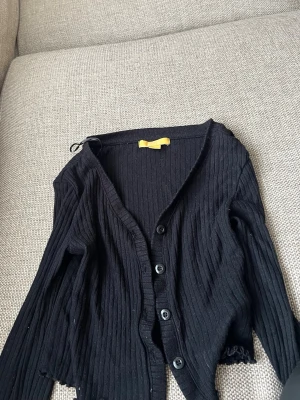 Svart ribbad kofta från Gina Tricot - Säljer en svart ribbad kofta från Gina Tricot med knappar framtill och lång ärm. Koftan har en smal passform och är tillverkad i ett mjukt, stretchigt material. Perfekt att slänga över en topp för en enkel och snygg look.