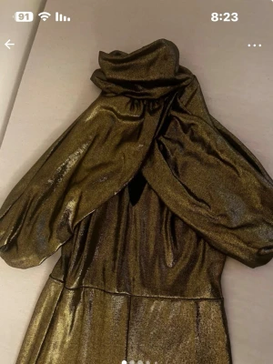 Glittrig guldklänning med unik hals - Säljer en glittrig guldklänning med hög hals och draperade, vida ärmar som ger en riktigt statement-look. Klänningen har en snygg v-ringning framtill och ett metalliskt skimmer i tyget. Perfekt för dig som vill sticka ut och glänsa på nästa event!