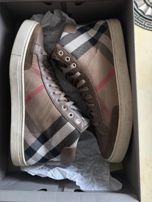 Burberry rutiga high top sneakers - Snygga high top sneakers från Burberry med klassiskt rutmönster i beige, svart, vitt och rött. Skorna har bruna mockadetaljer, vita sulor och är tillverkade i canvas och mocka. Perfekta för dig som vill sticka ut med en ikonisk design.