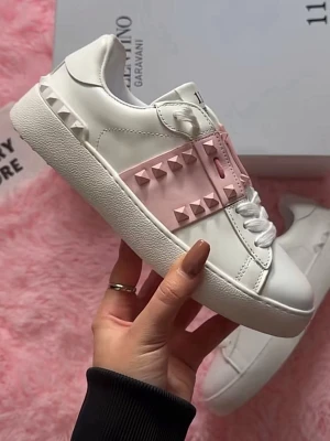 Valentino Rockstud sneakers vit/rosa - Snygga vita sneakers från Valentino med en bred rosa rem över snörningen dekorerad med ikoniska pyramidnitar. Skorna har chunky sula och är tillverkade i slätt läder. Perfekt för dig som vill sticka ut med en cool och trendig look.