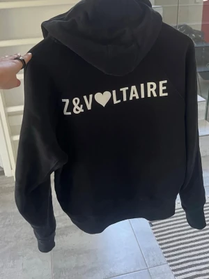 Svart hoodie från Zadig & Voltaire - Snygg svart hoodie från Zadig & Voltaire med stor vit logga och hjärtdetalj på ryggen. Klassisk modell med huva och känguruficka framtill. Perfekt för en chill och trendig look. Mjuk och skön bomullskänsla. Kvitto finns kvar, köpt från USA.