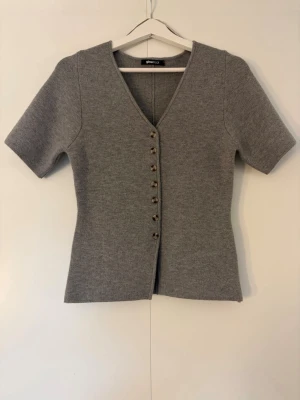 Grå kortärmad kofta från Gina Tricot - Stilren grå kofta från Gina Tricot med korta ärmar och v-ringning. Koftan har dekorativa knappar framtill och är gjord i ett mjukt, stickat material som ger en snygg passform. Ny utan prislapp. Oanvänd