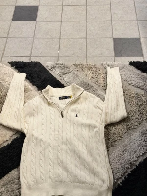 Vit kabelstickad tröja Polo Ralph Lauren - Kabelstickad vit tröja från Polo Ralph Lauren med halv dragkedja och klassisk marinblå logga broderad på bröstet. Tröjan har ribbade muddar och krage, perfekt för lager-på-lager och en clean look.