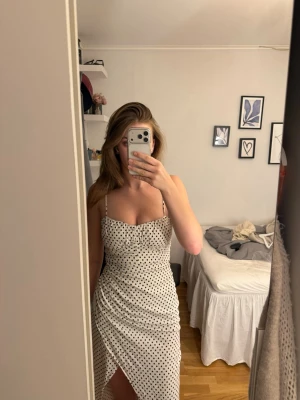Vit prickig klänning med slits - Supersnygg vit klänning med svarta prickar och smala axelband. Klänningen har en figurnära passform, omlottkänsla och en slits framtill som ger en trendig look. Perfekt för dig som gillar en feminin och stilren stil. Skulle säga att den är mindre än L. 