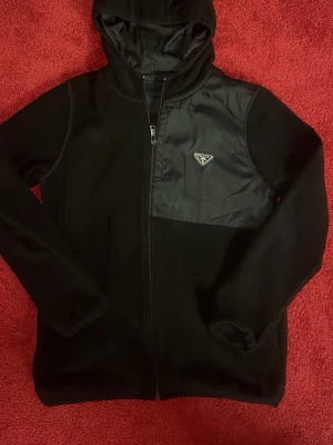 Fleecejacka - Snygg svart hoodie från Prada med dragkedja framtill och en cool detalj i form av en blank bröstficka med logotyp. Tillverkad i mjukt material och har en klassisk huva. Perfekt för dig som vill ha en stilren och exklusiv look.