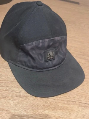 Svart keps från Hugo med mönster - Snygg svart keps från Hugo med ett diskret mönster på frontpanelen och en metallpatch med logga. Klassisk böjd skärm och justerbar baksida. Perfekt accessoar för en trendig streetwear-look.