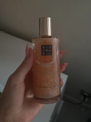 Rituals Shimmering Body olja - Lyxig kroppsmist från Rituals med skimrande effekt. Flaskan är genomskinlig och innehåller en gyllene vätska med glitterpartiklar. Perfekt för att ge huden en fräsch glow och doft. Kommer i en elegant flaska med guldfärgat lock.