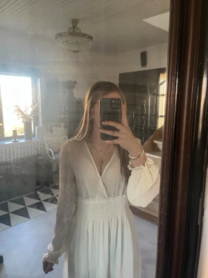Vit kortklänning med v-ringning - Säljer en vit kortklänning med v-ringning och långa, transparenta ärmar. Klänningen har smock i midjan och volangdetaljer vid ärmslut. Perfekt för skolavslutning,student och midsommar!